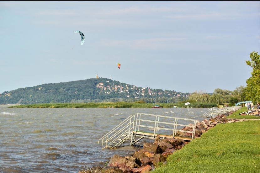 Szörfösök és sárkányszörfösök a balatonfenyvesi parton, háttérben a Fonyódi-heggyel